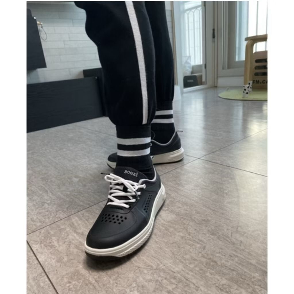 Person wearing black golf sneakers with white soles on a tiled floor.
