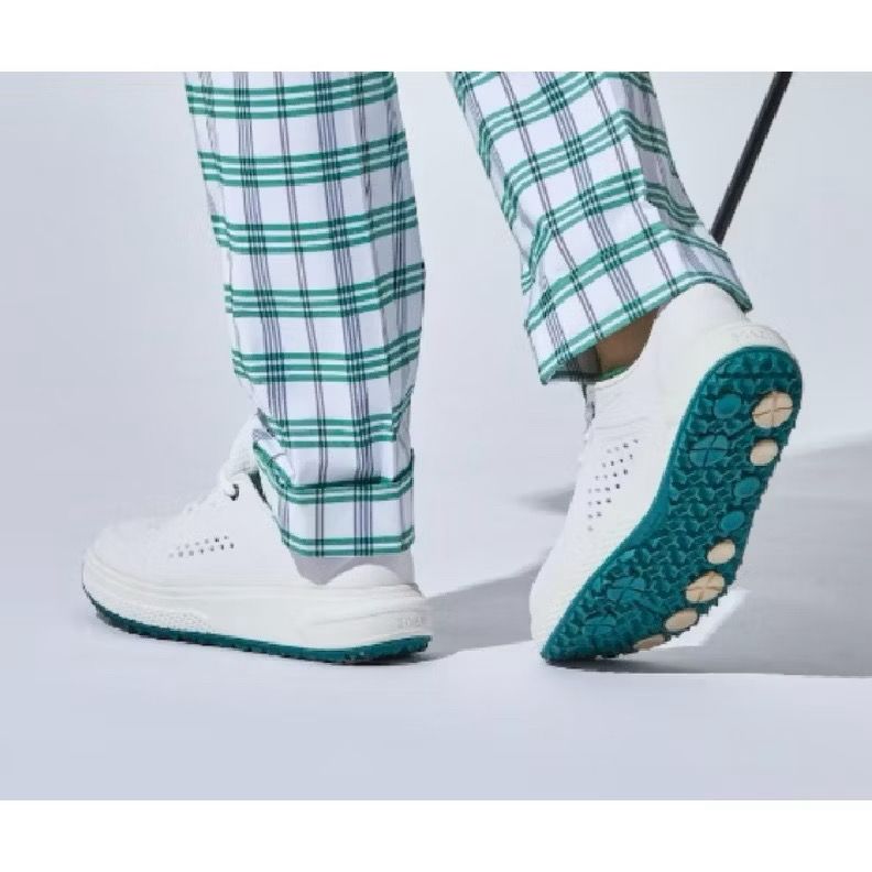 White golf sneakers with green sole on checkered pants against a light gray background